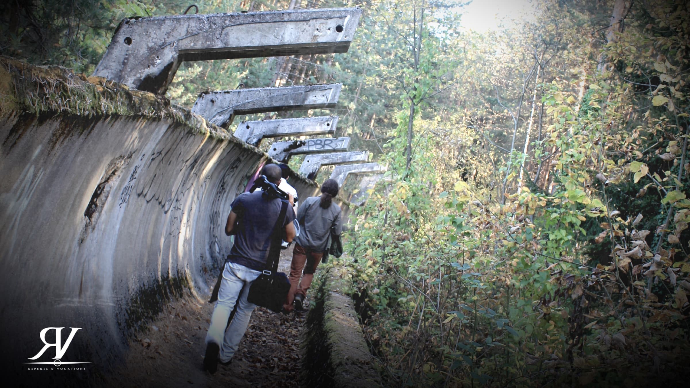 Deux membres de l’équipe, caméra à l’épaule et sac à la main, remontent une piste de bobsleigh en béton envahie par la forêt.