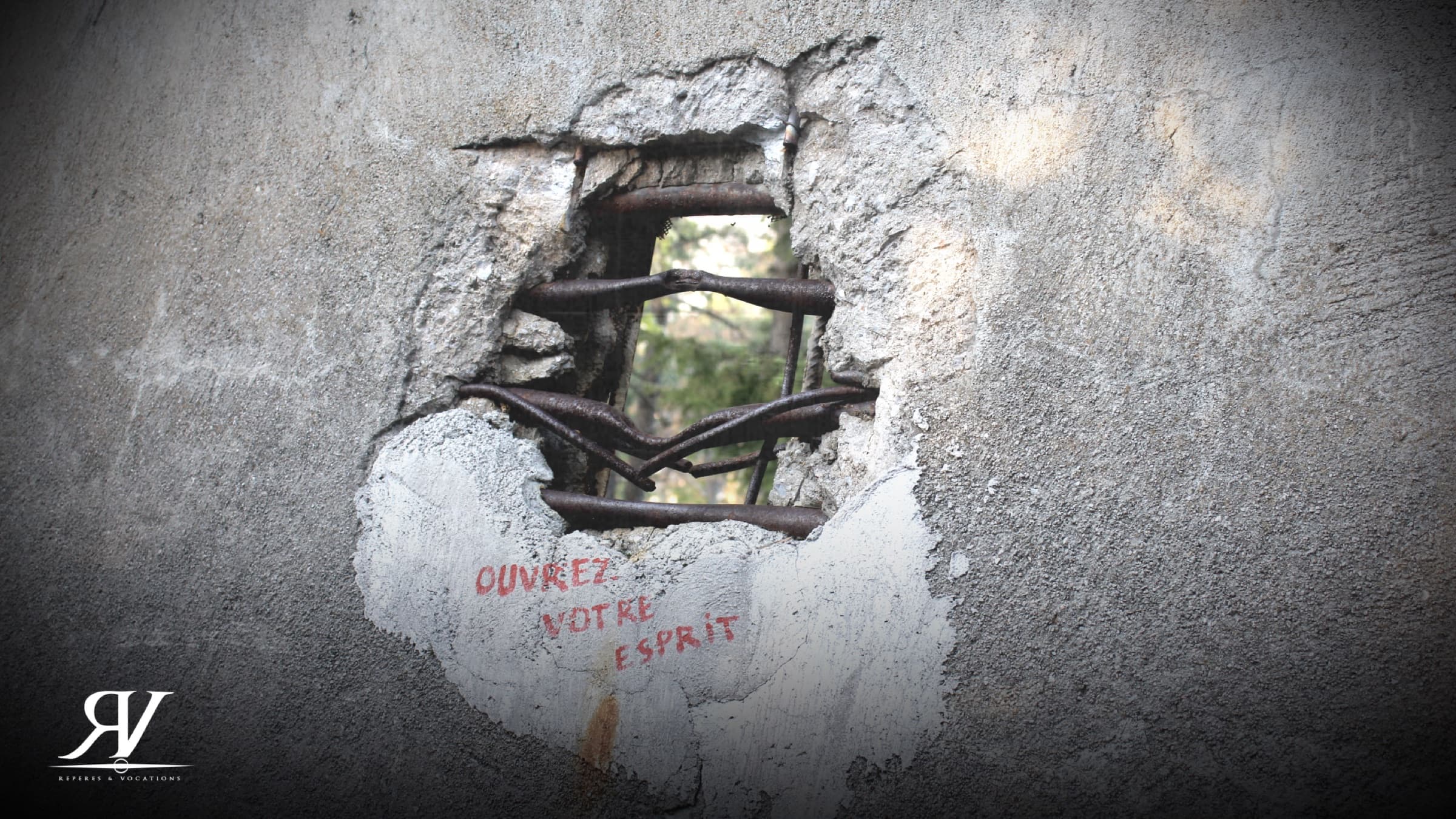 Trou d’obus dans un mur de béton, les fers à béton tordus vers l’extérieur ; en dessous, trois mots peints en rouge : « Ouvrez votre esprit ».