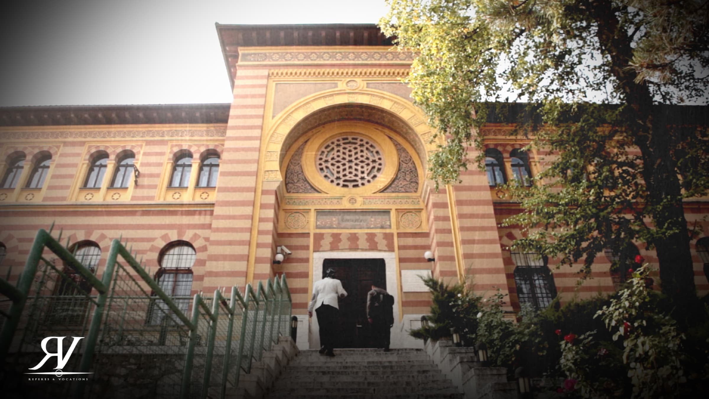 Façade en bandes de pierre ocre et rose d’un bâtiment de style ottoman revisité, grand portail en arc surmonté d’une rosace ; deux personnes gravissent l’escalier d’entrée.