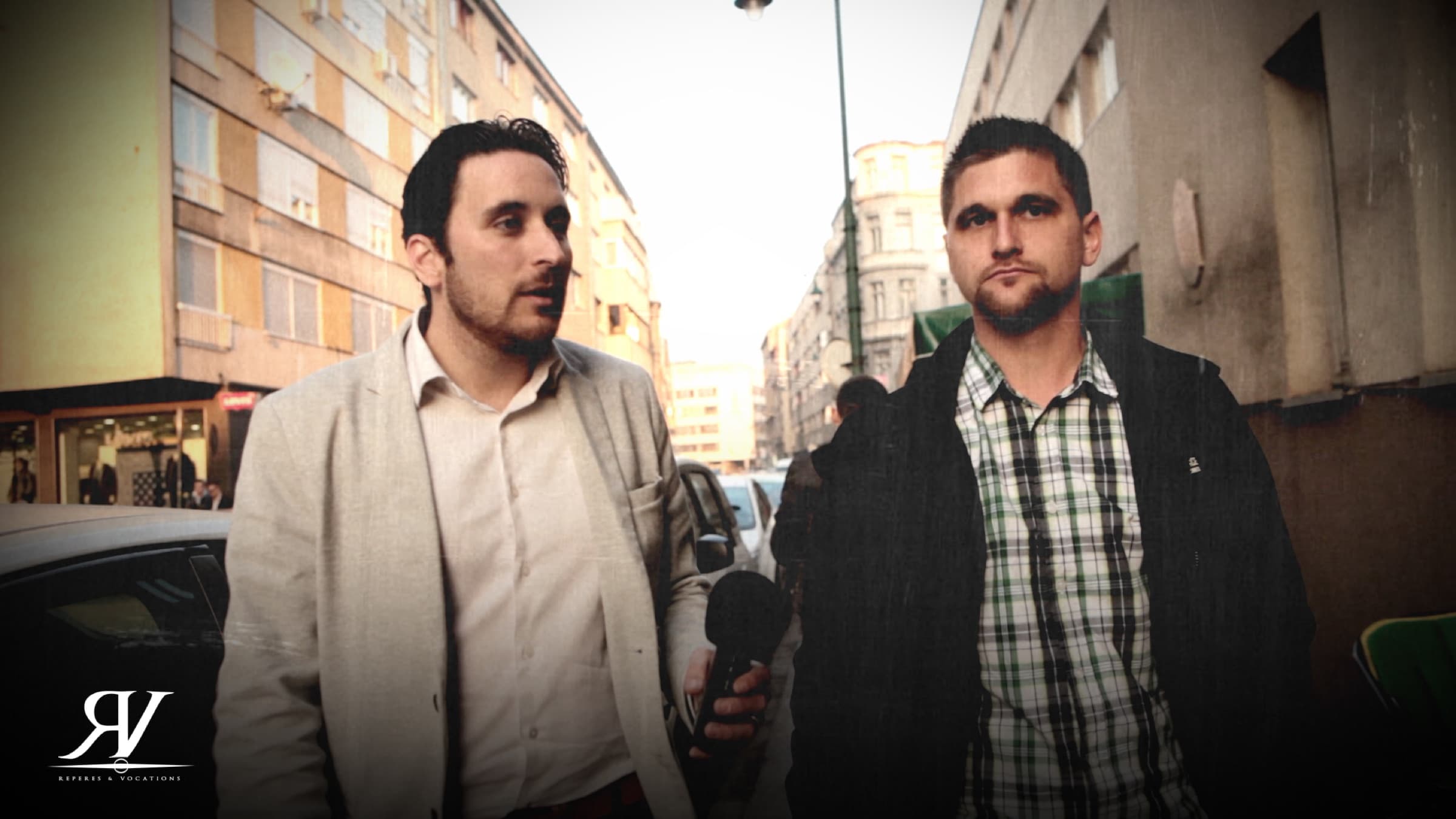 Deux hommes marchent côte à côte dans une rue commerçante ; celui de gauche, en veste claire, tient un micro-main baissé le long du corps.