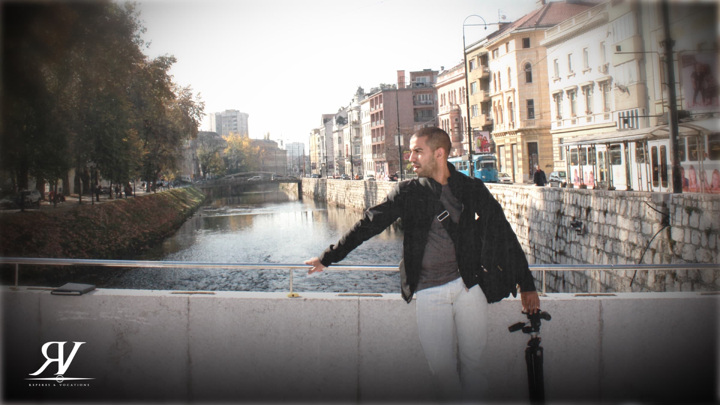Un homme en veste noire, un pied de caméra à la main, appuyé au parapet d’un quai ; derrière lui, la rivière encaissée et les façades austro-hongroises de Sarajevo.