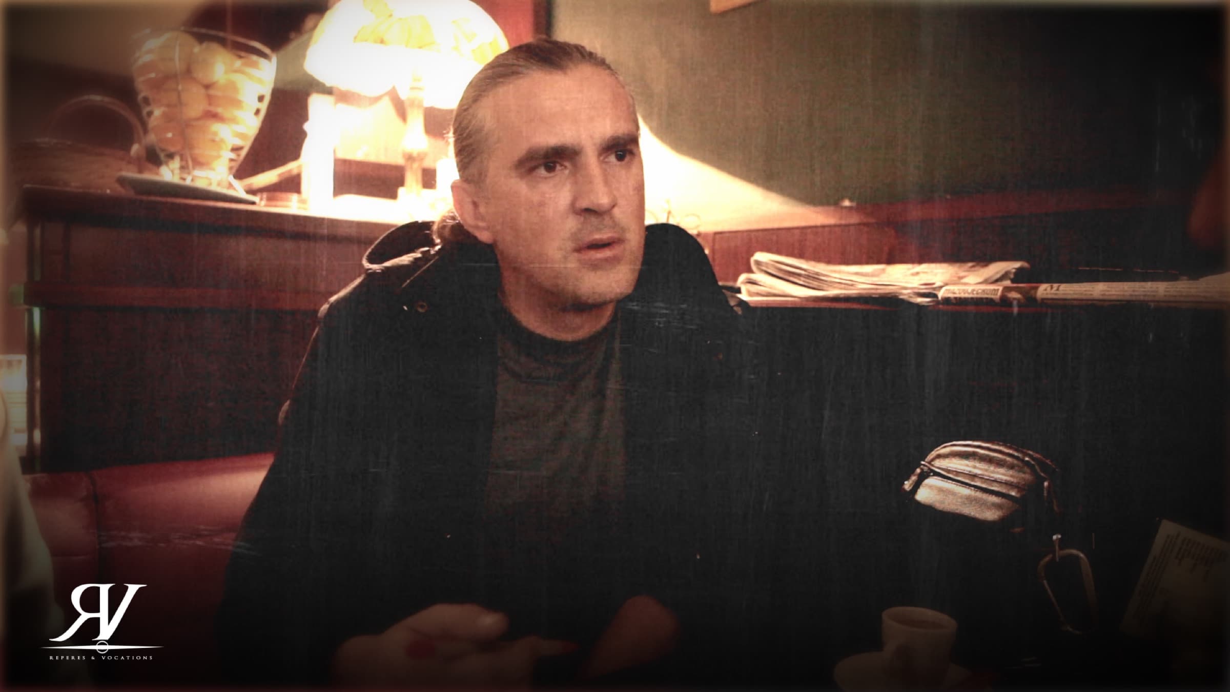 Portrait d’un homme aux cheveux tirés en arrière, assis dans une salle de café en bois sombre, en train de parler à quelqu’un hors champ.