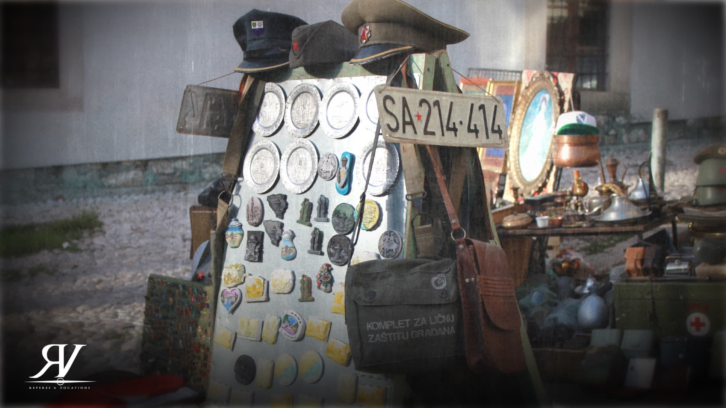 Étal de brocante en plein air : panneau couvert de médailles et d’insignes militaires, casquettes d’uniforme, sacoche kaki et vieille plaque d’immatriculation de Sarajevo.