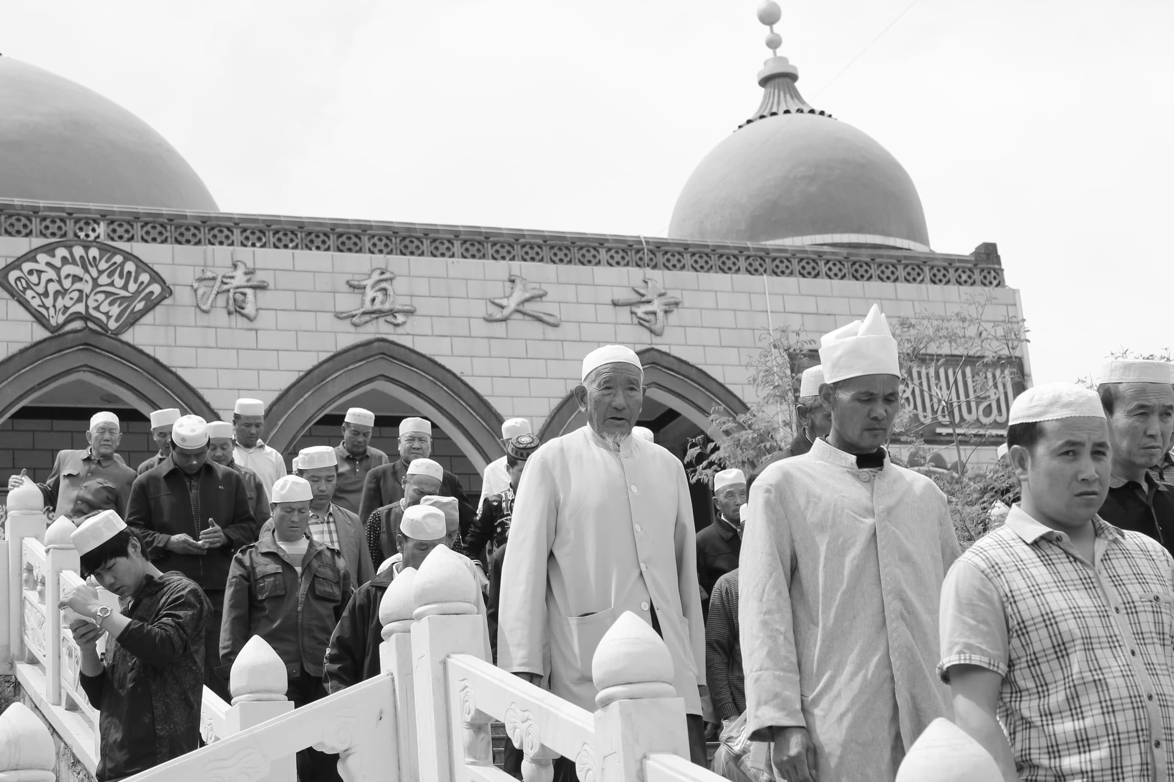 Foule d’hommes en calot blanc massés devant le portail d’une mosquée à coupoles, sous une inscription en caractères chinois et en arabe.