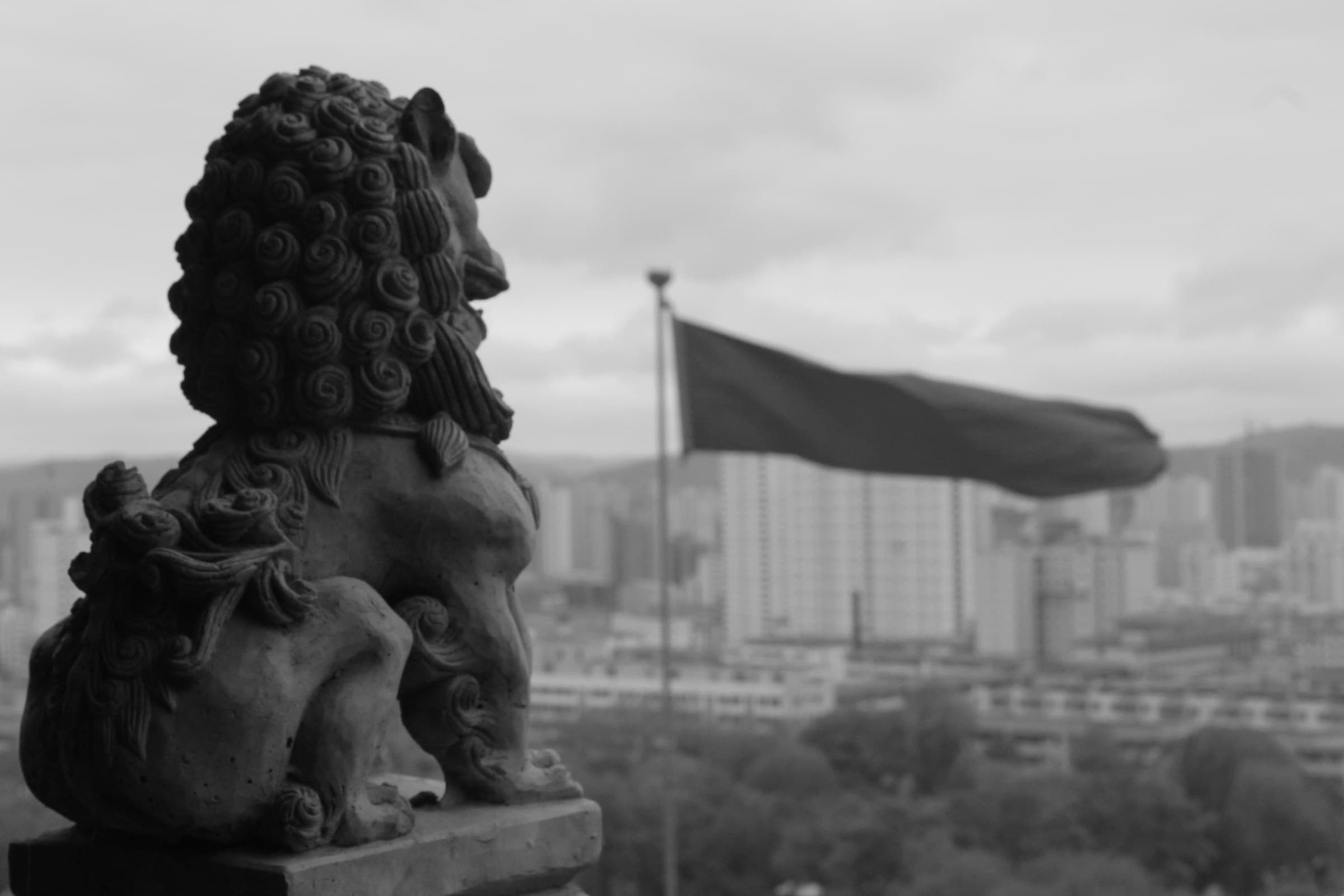 Lion de pierre monumental dressé au-dessus de la ville ; derrière lui, un grand drapeau claque au vent au-dessus d’un océan d’immeubles.