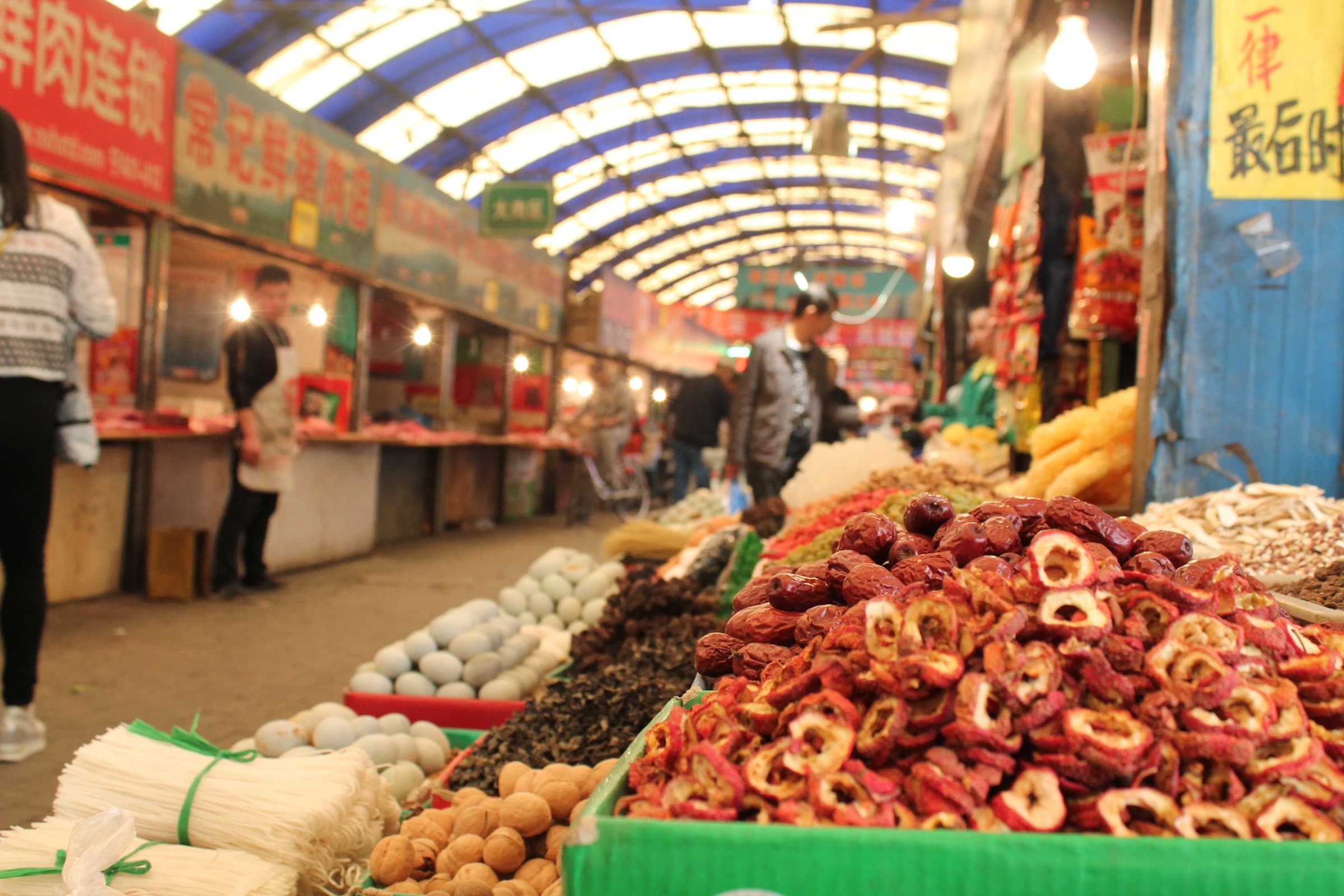 Longue allée de marché couvert : bacs débordants de piments séchés, de fruits confits et de racines, sous une verrière en berceau.