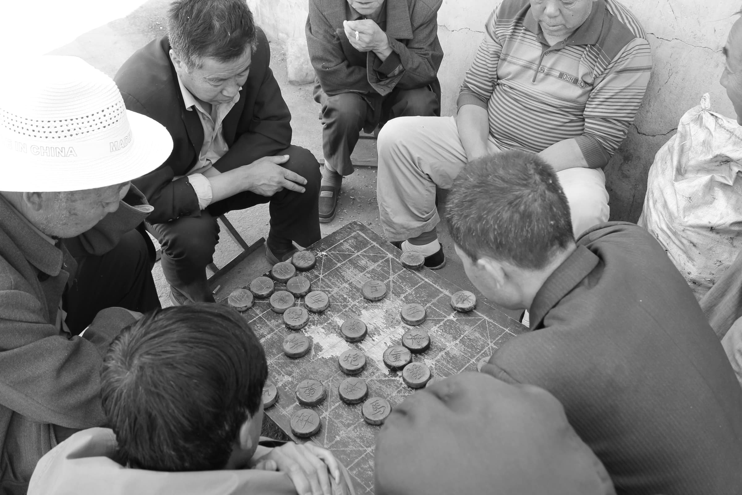 Petit attroupement d’hommes penchés sur un damier posé à même une table basse, dans la rue ; l’un d’eux porte un chapeau de paille clair.