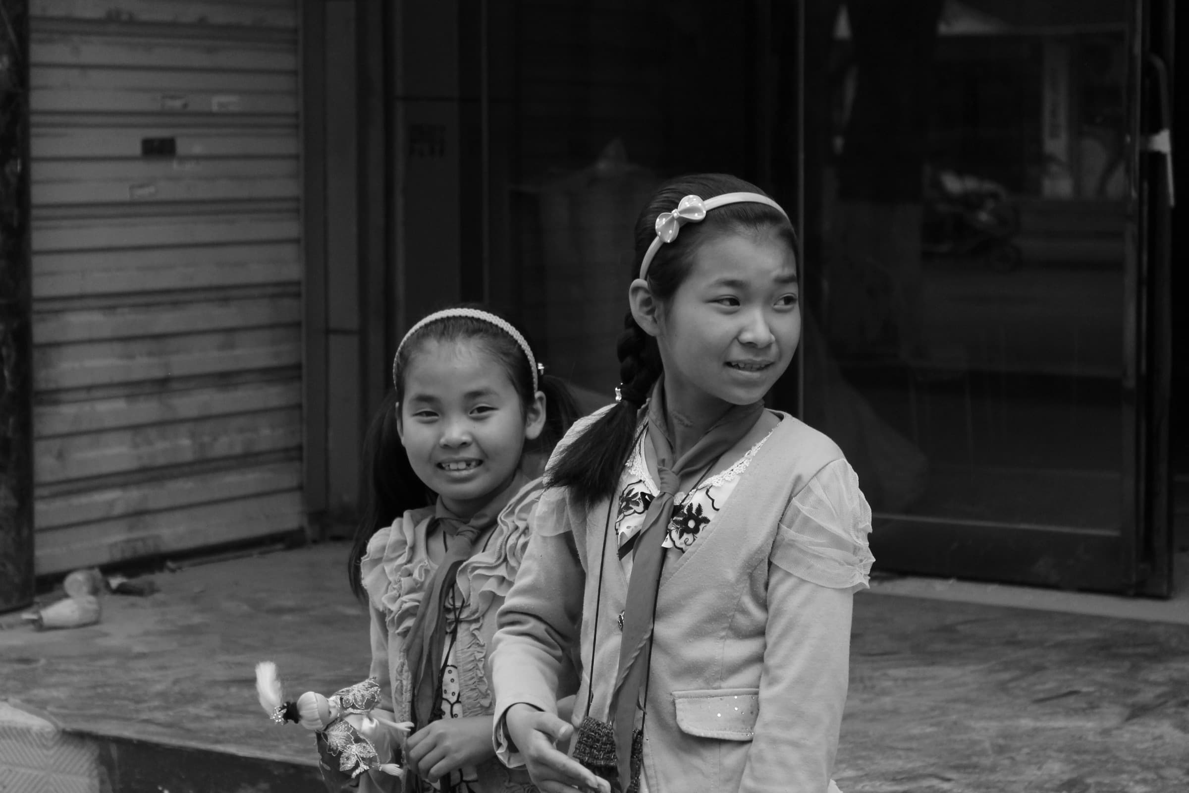 Deux fillettes qui se ressemblent trait pour trait, en veste claire et coiffure identique, sourient côte à côte devant un rideau de fer.
