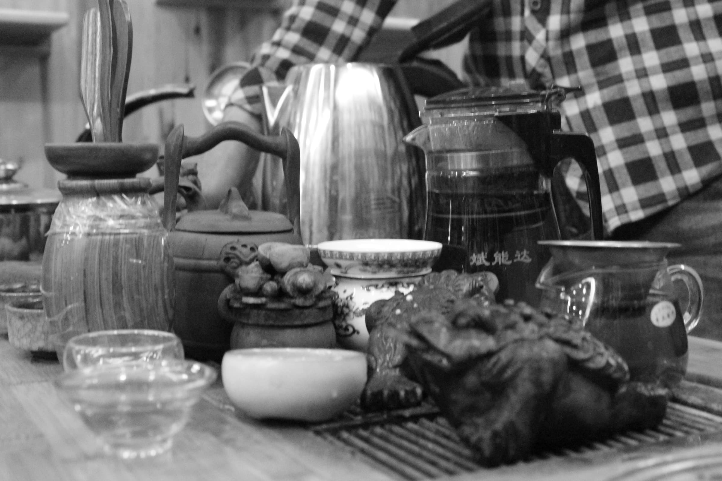 Sur un plateau de bois, une théière de métal, des bocaux de verre et de petites tasses entourent un amas de feuilles ; une main en chemise à carreaux verse l’eau.