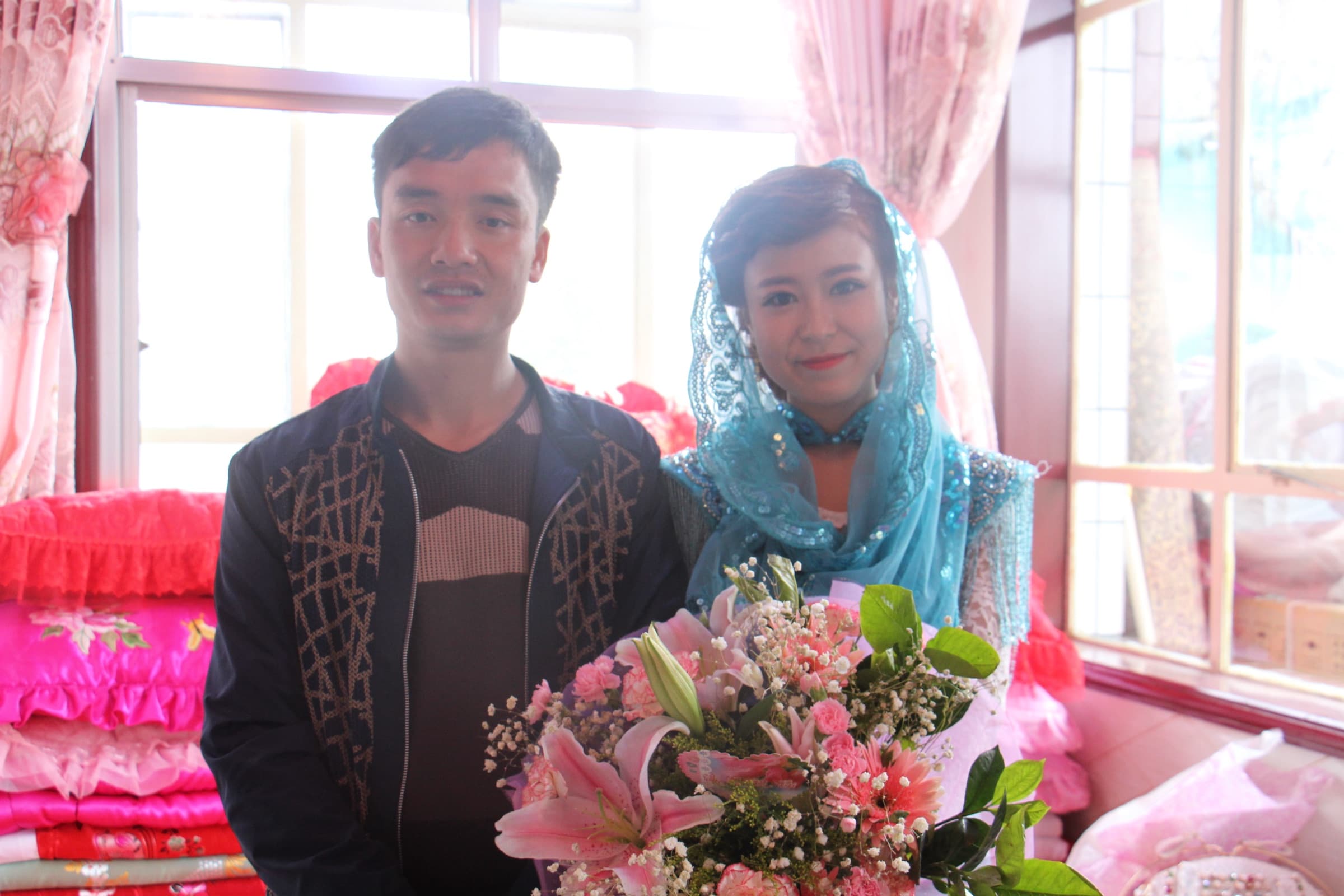 Un jeune couple pose côte à côte, bouquets à la main, devant une tenture et des fleurs de papier rose ; la femme porte un voile de tulle vert.