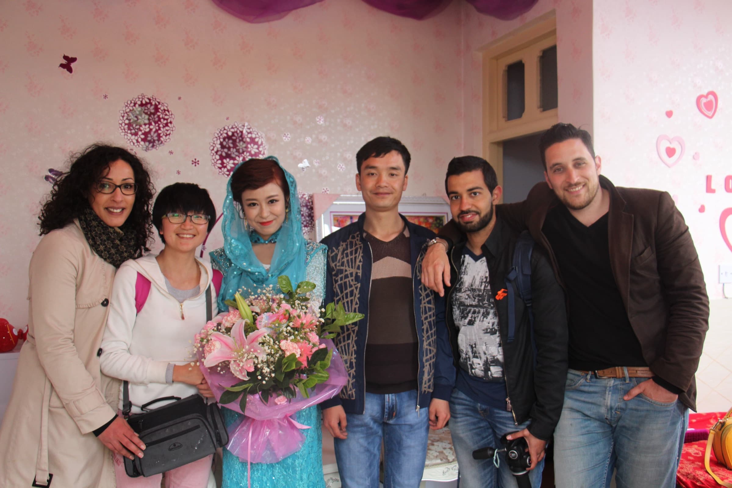 Six personnes posent serrées devant un mur décoré de fleurs roses : les membres de l’équipe entourent la famille et la jeune femme qui tient un bouquet.